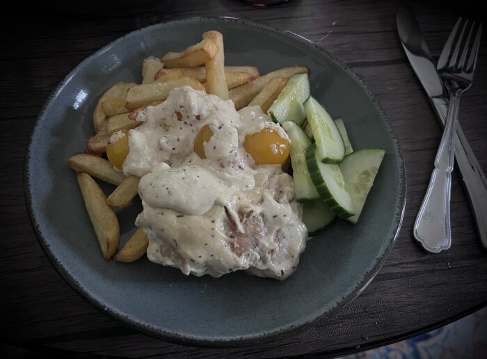 Tallrik med pommes frites, gurkskivor och kyckling i krämig sås toppad med gula tomater.