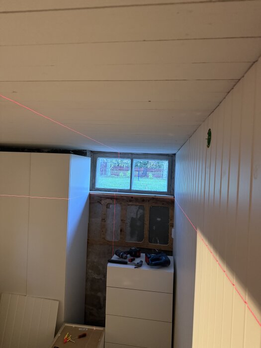 Källare med plywoodram på väggen för att fästa panelskivor, laserlinjer markerar ramar, fönster ovan, verktyg på byrå.