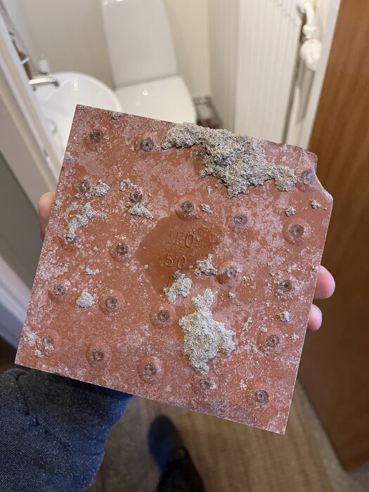 Närbild av en fyrkantig, rödbrun klinkerplatta med cementrester och skador, hålls i handen i ett badrum.