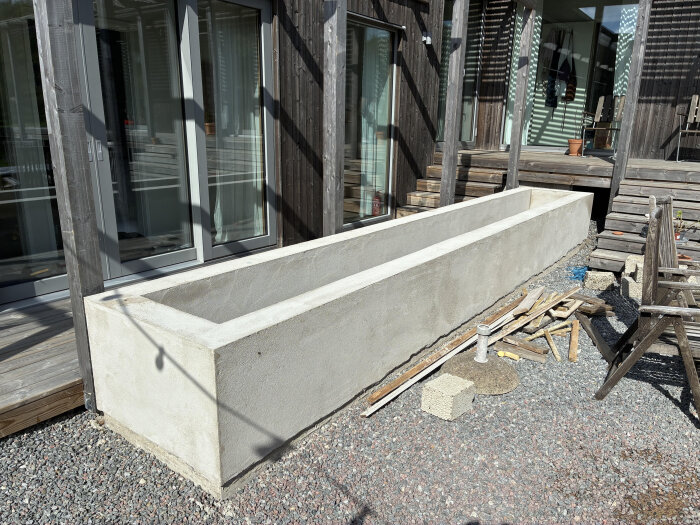 Murad och putsad blomlåda i lecablock med omkringliggande byggmaterial, placerad på grus bredvid en uteplats och husfasad.
