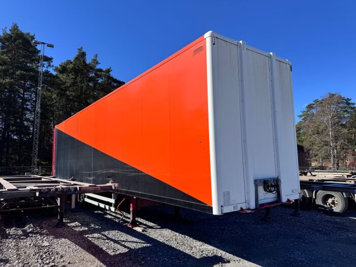 En stor orange och vit trailer parkerad utomhus på grus, omgiven av skog.