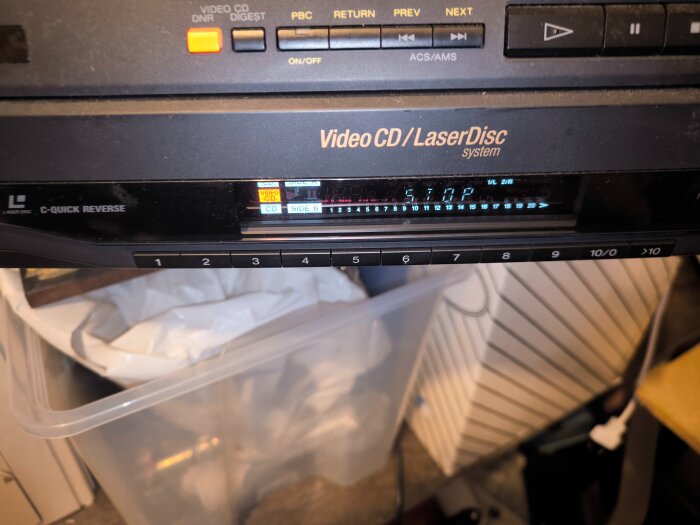 Laserdisc-spelare med kontrollpanel och display som visar "STOP". Spelaren står ovanpå en plastlåda i ett förråd.