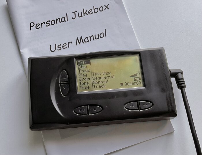 Bärbar jukebox med skärm och knappar, kopplad med sladd. Manual i bakgrunden med texten "Personal Jukebox User Manual".