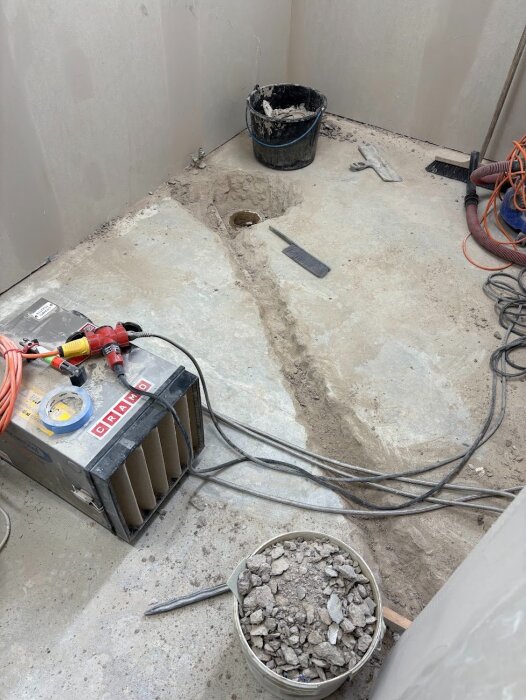 Byggarbetsplats med bilning på golvet, synliga rör, en svart hink med spillmaterial och verktyg spridda på golvytan.