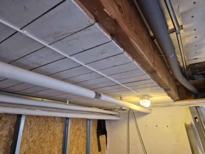 Gammalt träbjälktak med rör och slangar längs väggen i ett renoveringsprojekt, elektrisk belysning synlig.