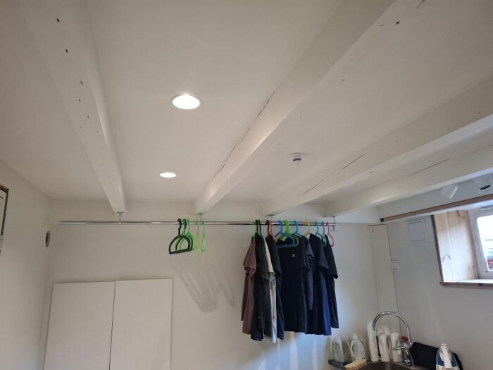 Vita bjälkar i tak med klädhängare och kläder i ett rum under renovering, med mörka väggskåp och fönster.