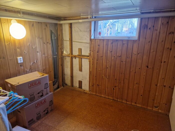 Renoverat rum med träpanel på väggarna, småfönster och uppackade flyttlådor på golvet. Synliga rör längs tak och vägg.