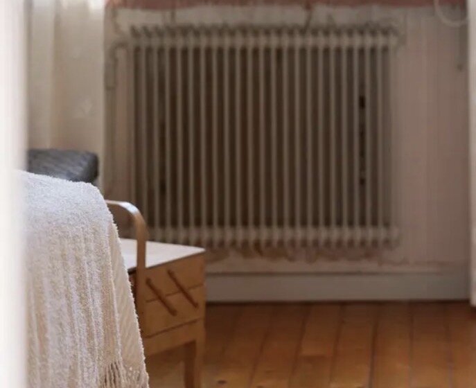 Radiator med vit ribbad design i träbeklätt rum, bredvid en stol med vit filt på.