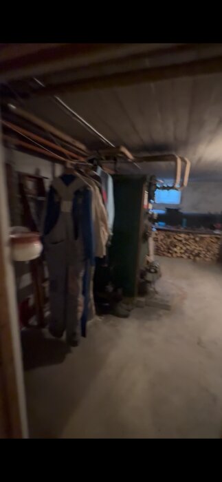 Källare med pannrum; grönt värmesystem, vedstapel, upphängda overaller och rör i taket. Mörkt rum med grått betonggolv.
