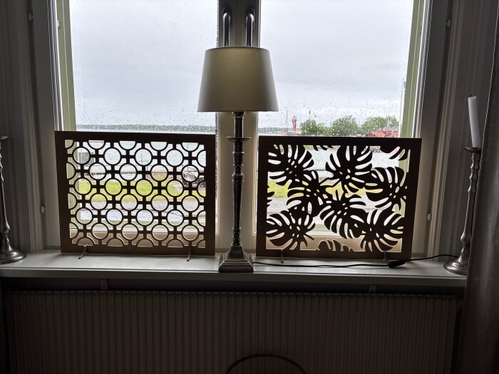 Träpaneler med utskurna mönster av cirklar och lövverk står på en fönsterbräda, belyst av en lampa.