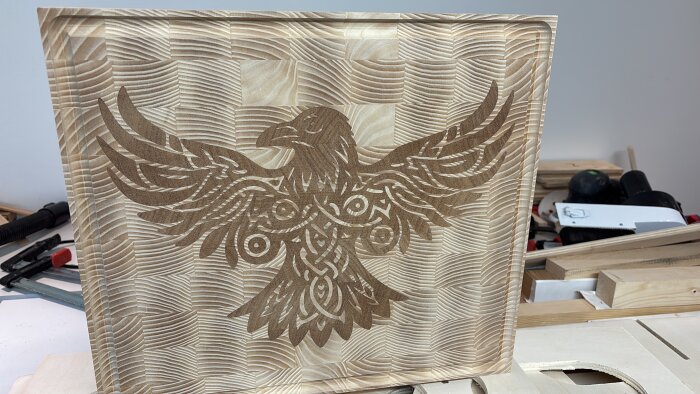 Träsnidat mönster av en fågel med utbredda vingar på en träpanel, skapad med en CNC-maskin. Bakgrunden visar verktyg och träskivor.