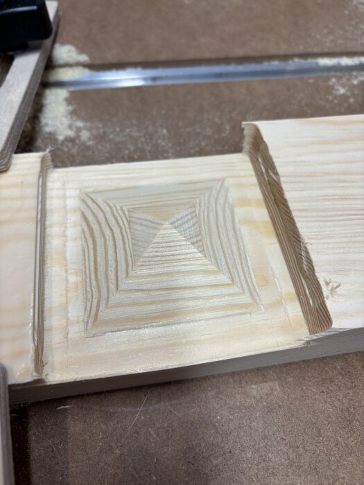 Närbild på träbit med ett pyramidformat mönster som skapats av en CNC-maskin, med synligt sågspån på arbetsytan.