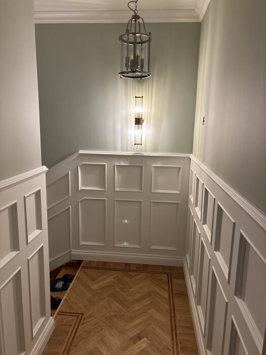 Trappa med väggpaneler och snyggt trägolv i fiskbensmönster, upplyst av en taklampa och vägglampor.