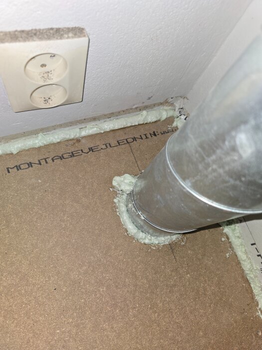 Skum sprutat mellan vägg och spånplattor runt ett ventilationsrör på ett nylagt nivellgolv, med ett vägguttag synligt i bakgrunden.