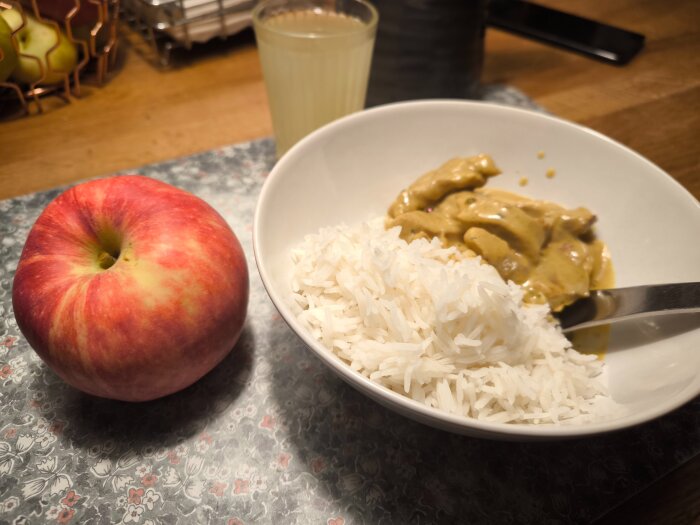 En tallrik med basmatiris och fläskfilé i currysås bredvid ett stort Amorosa-äpple på ett blommigt underlägg.