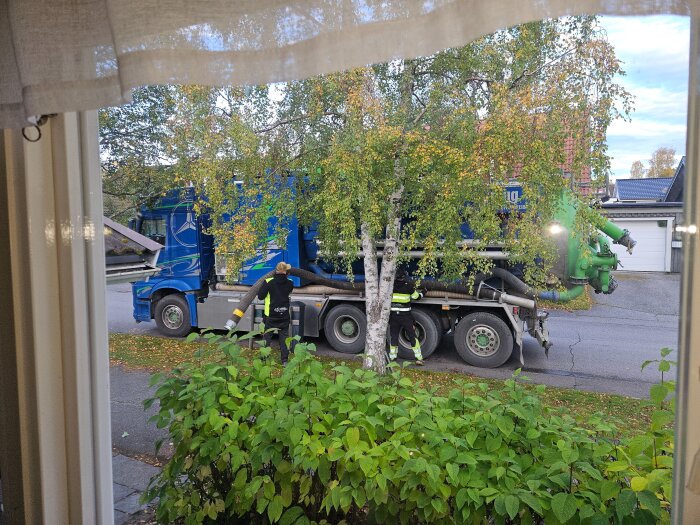 Blå och grön dammsugarbil parkerad på gatan, omgiven av träd och buskar, med en person som står vid fordonet.