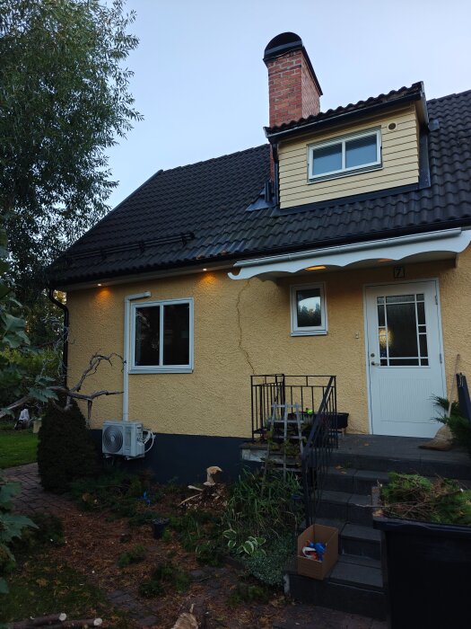 Ett gult hus med svart tak och en skorsten. En entré med trappa och växter samt en luftvärmepump syns framför huset.