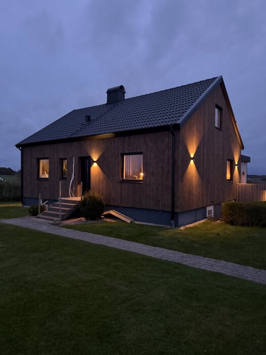 50-tals hus med ny fasad och belysning i skymning. Gräsmatta och stenlagd gång i förgrunden.