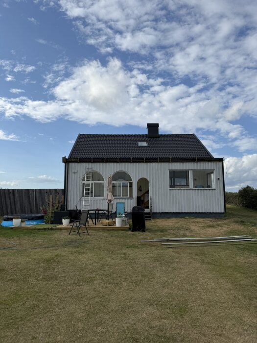 50-tals hus med ny fasad och tak, sett framifrån med trädgårdsmöbler och en stege, under en blå himmel med moln.