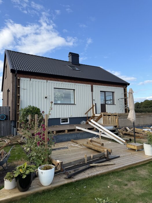 50-tals hus med ny fasad och pågående rivning av uterum på träaltan, för att påbörja poolbygge. Gräs och växter i förgrunden.