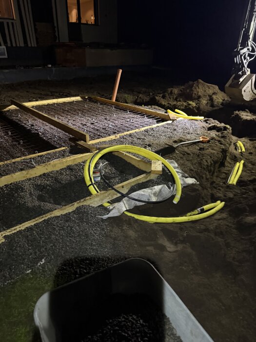 Form och golv för betongplatta under byggnation på natten, med synliga kablar och verktyg, bredvid en grävmaskin.