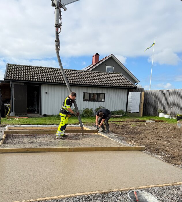 Två personer gjuter en betongplatta med hjälp av en betongpump vid ett hus, förbereder teknikrum under altanbygge.