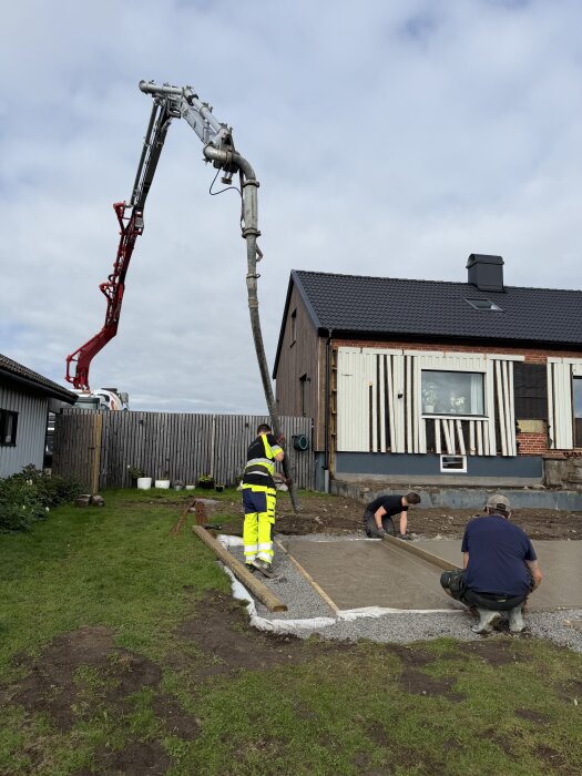 Betongplatta gjuts nära ett hus, tre personer arbetar med ytan. En betongpump används, och husets fasad renoveras delvis i bakgrunden.