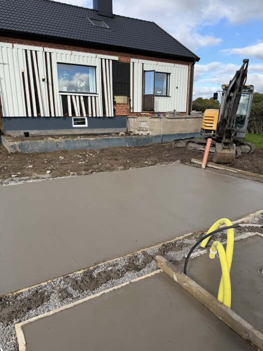 En nygjuten betongplatta framför ett hus med delvis riven panel, en grävmaskin och gula slangar i förgrunden.