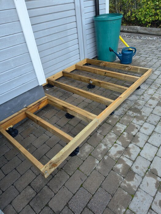 Träram till förråd byggs mot huset, med fötter i marksten. Grön soptunna och blå vattenkanna syns i bakgrunden.