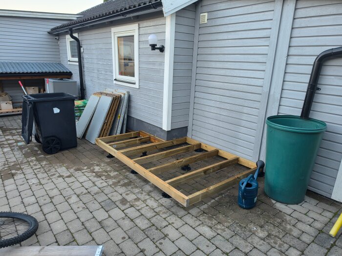 Träram för förrådsgrund byggd intill ett hus med grå träpanel, omgiven av sopkärl, en grön tunna och en blå vattenkanna.