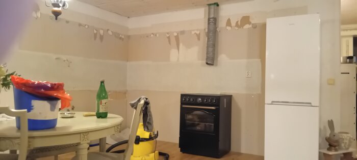 Kök med borttagna skåp och vitvaror, synlig spis och kylskåp, bord med flaska och hink, redo för renovering.