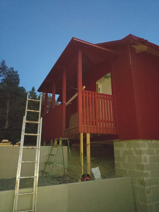 Rött trähus under konstruktion på en slutning, stege och byggmaterial i förgrunden, kvällstid.
