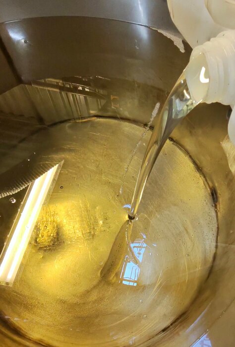 Vätska från en flaska hälls i en metalltunna, reflekterar ljus från en taklampa.