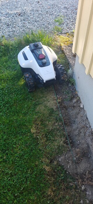 Robotgräsklippare intill en husvägg, med en grusuppfart i bakgrunden.