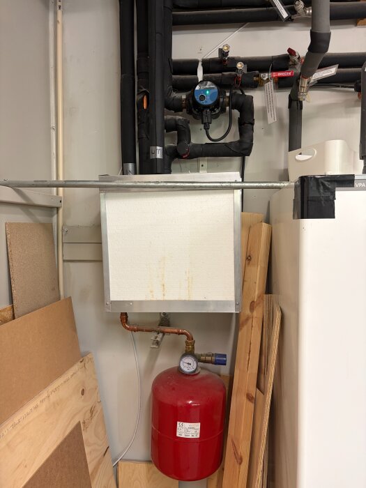 Värmesystem med rör, expansionskärl och manometer i ett hus, omgiven av träpanel och vägg.