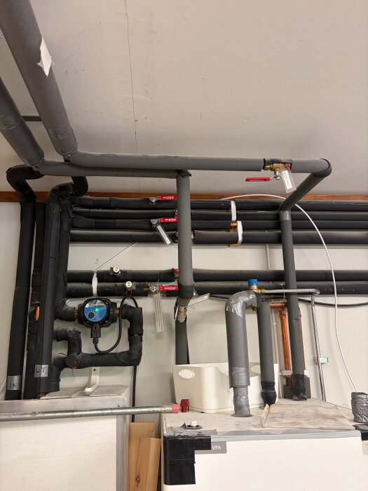 Rörsystem och värmepump i en källare, visar installationen av fjärrvärmeanläggning med ventiler och isolerade rör för varmvatten och uppvärmning.