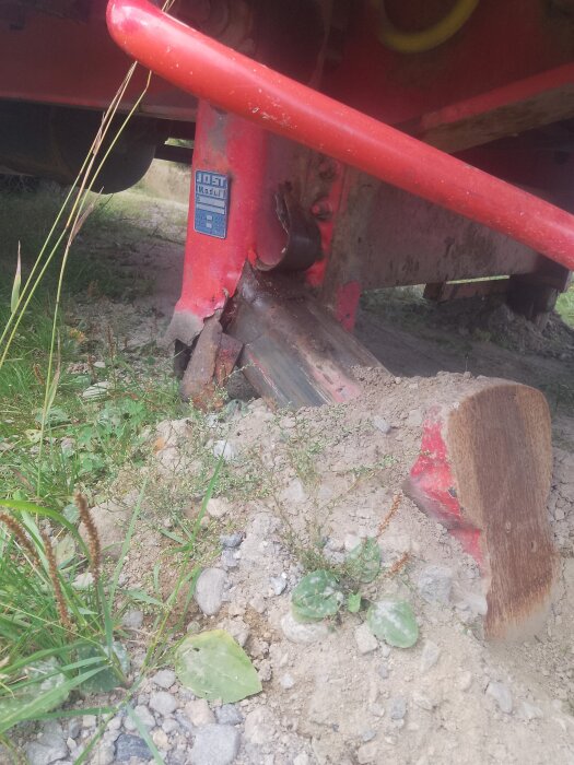 Vikt stödben på röd släpvagn vilar på ojämn mark omgiven av gräs och stenar, vilket försvårar transport och kräver reparation.