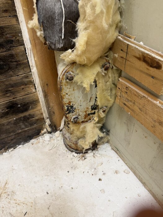 Ett gammalt metallrör i ett badrumshörn täckt med isolering och rost, omgivet av träpaneler och betongrester på golvet.