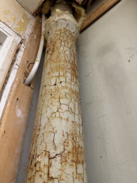 Rostigt rör i hörn av badrum under renovering, går rakt upp mot papptak, omgiven av trä och ledningar. Rörets funktion är okänd.
