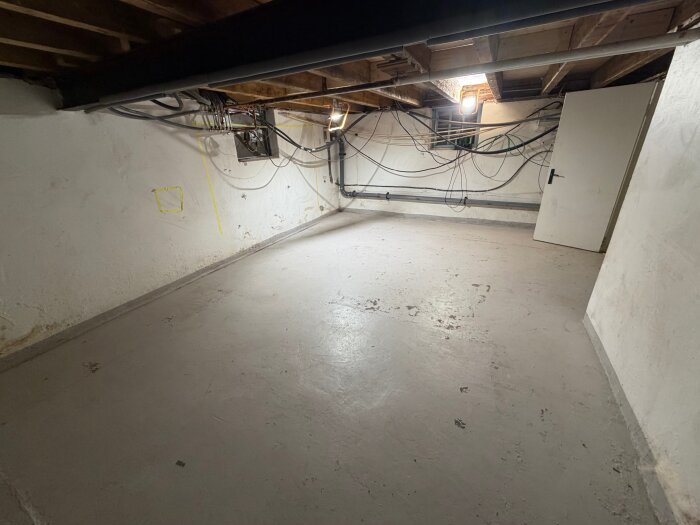 Källare med betonggolv, vita väggar och synliga takbjälkar. Elektriska ledningar och en strålkastare är monterade på väggen.