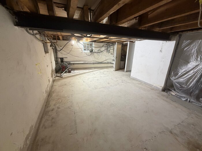 Källarutrymme under renovering med blottat tak, synliga rör och kablar. Betonggolvet är sprucket och en dörr leder till ett annat rum.