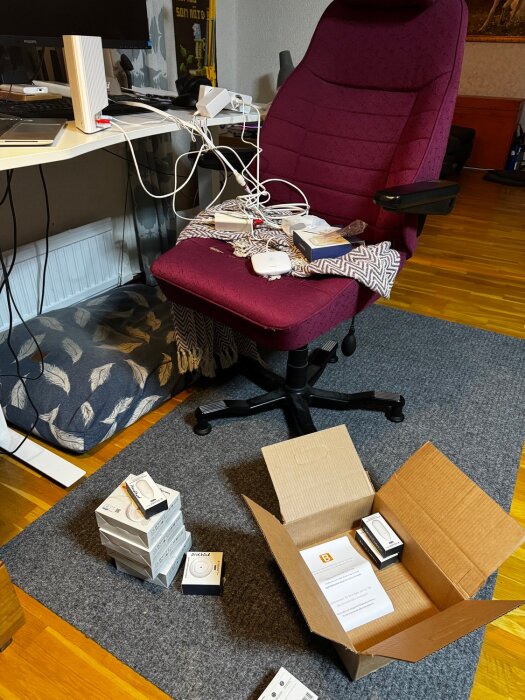 Arbetsrum med skrivbordsstol, dator, sladdar och en öppen kartong på golvet bredvid flera smarta hem-enheter i deras förpackningar.