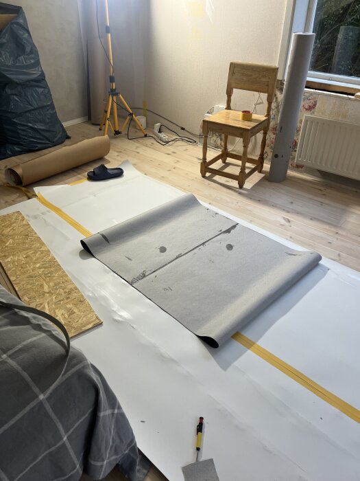 Rum under renovering med lumppapp på golvet, byggmaterial och verktyg utspridda; trätstol står vid väggen nära ett fönster.