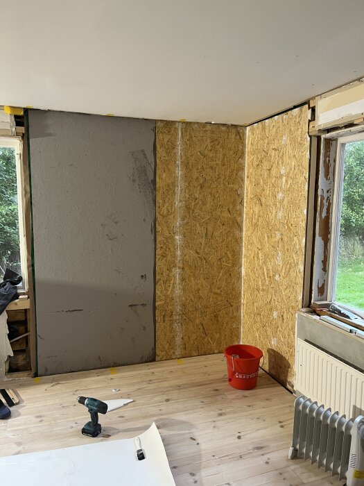 Rum under renovering med lumppapp och OSB-skivor på väggen, borrmaskin och röd hink på golvet, element och fönsterram utan glas.