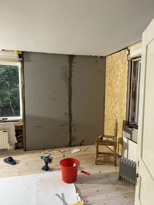 Rum under renovering med lumppapp på vägg, borrmaskin och röd hink på golvet, samt en fönsteröppning under arbete.