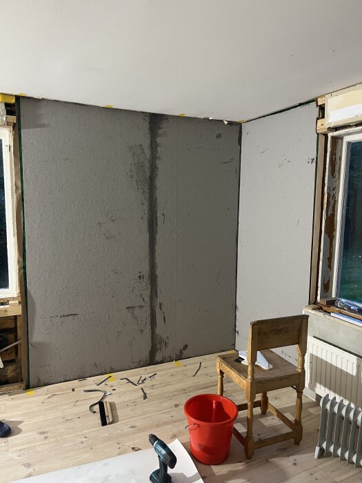 Vägg med lumppapp uppsatt, borrmaskin och röd hink på golvet, stol vid fönster, radiator vid golvet.
