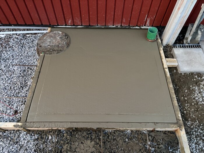 Betongplatta i träform mot röd vägg, med en cirkulär yta och ett grönt rör. Omgiven av grus och jord.