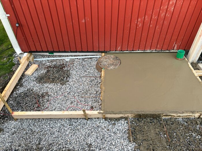 Betongplatta under konstruktion bredvid en röd vägg, omgiven av träformar och grus på byggarbetsplats.