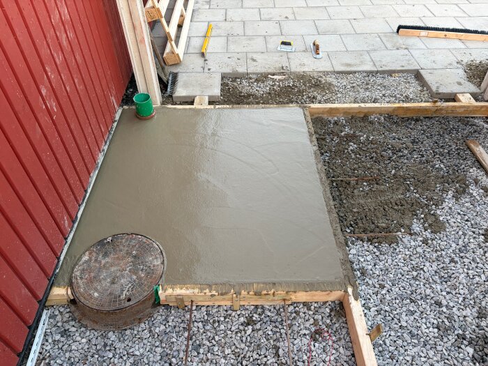 Nygjuten betongplatta i en träform intill röd byggnad, omgiven av grus och verktyg.