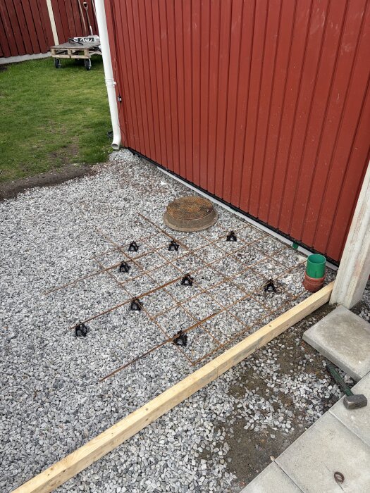Armeringsjärn på grusad yta framför röd fasad, preliminär betongbeläggning för byggprojekt, träplankor och avloppslock synliga.